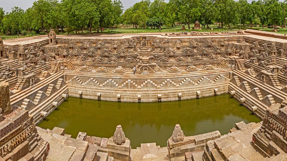 An ancient stone amphitheater with a central water body, surrounded by tiered stone seating and intricate carvings.