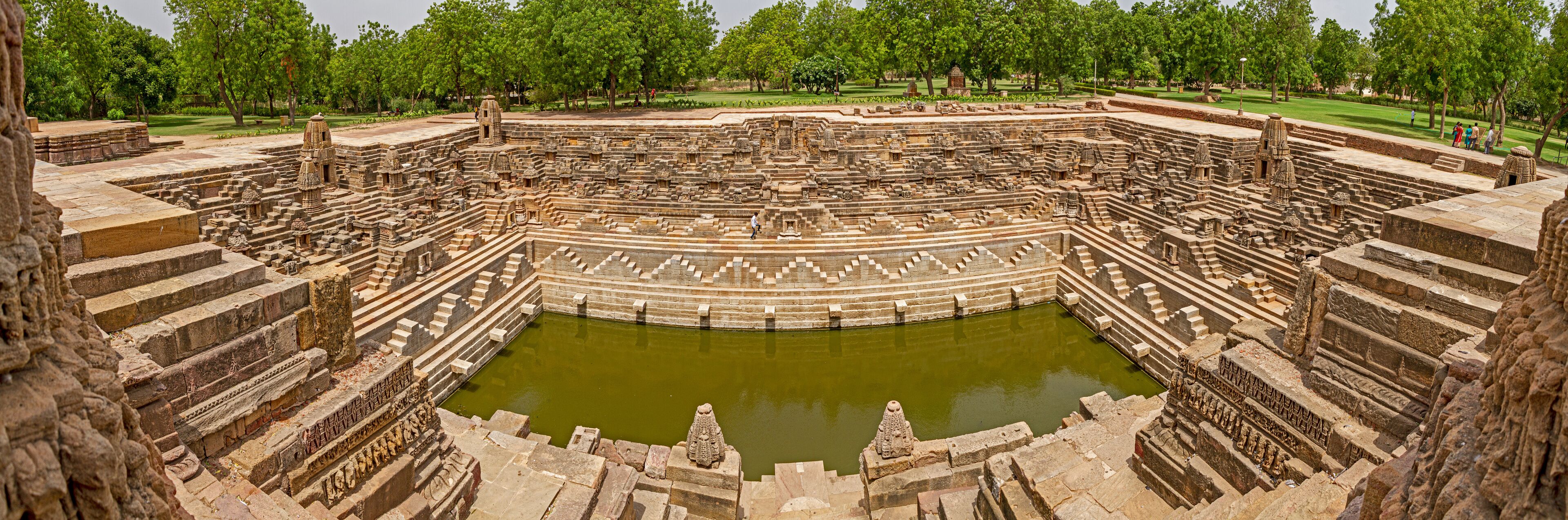An ancient stone amphitheater with a central water body, surrounded by tiered stone seating and intricate carvings.