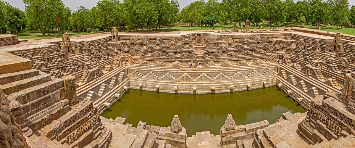 An ancient stone amphitheater with a central water body, surrounded by tiered stone seating and intricate carvings.