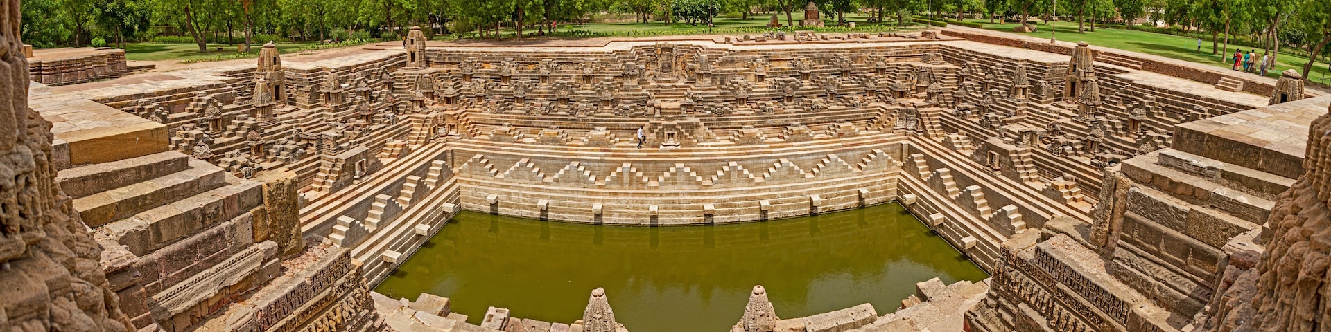 An ancient stone amphitheater with a central water body, surrounded by tiered stone seating and intricate carvings.