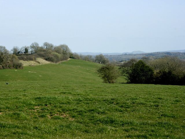 A green field with a hill, trees, and a distant view of hills.