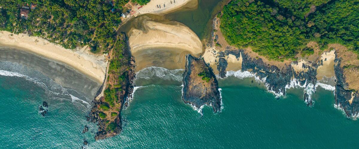 An aerial view of a coastal area with a sandy beach, rocky outcrops, and dense greenery.