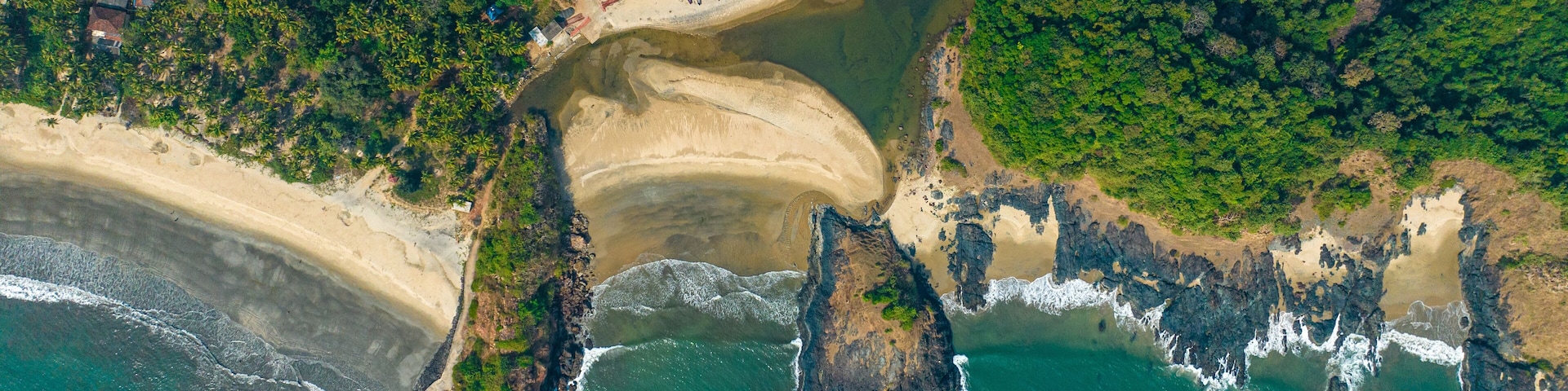 An aerial view of a coastal area with a sandy beach, rocky outcrops, and dense greenery.