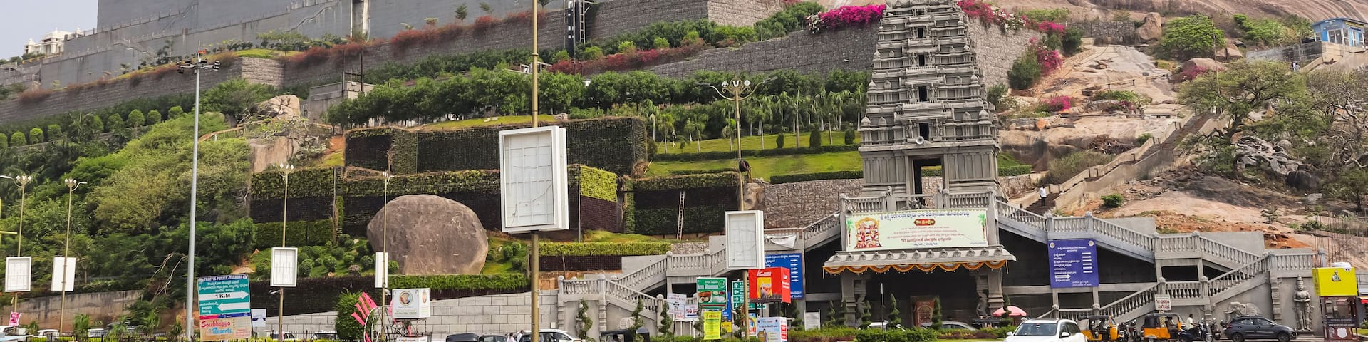 A large fort-like structure with a temple on a hill, surrounded by greenery and a busy street with vehicles.