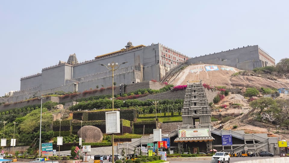 A large fort-like structure with a temple on a hill, surrounded by greenery and a busy street with vehicles.