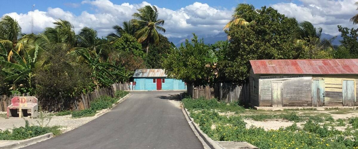 A rural road flanked by greenery and a small blue building with a red door.