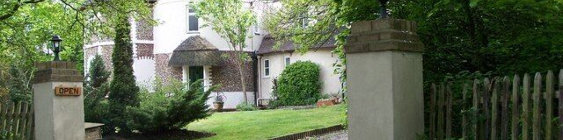 Un camino de grava que conduce a una casa blanca con chimenea de ladrillo, rodeada de abundante vegetación y una cerca de madera.