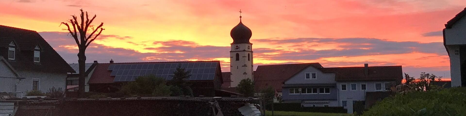 A church with a clock tower, surrounded by houses and a grassy field during sunset.