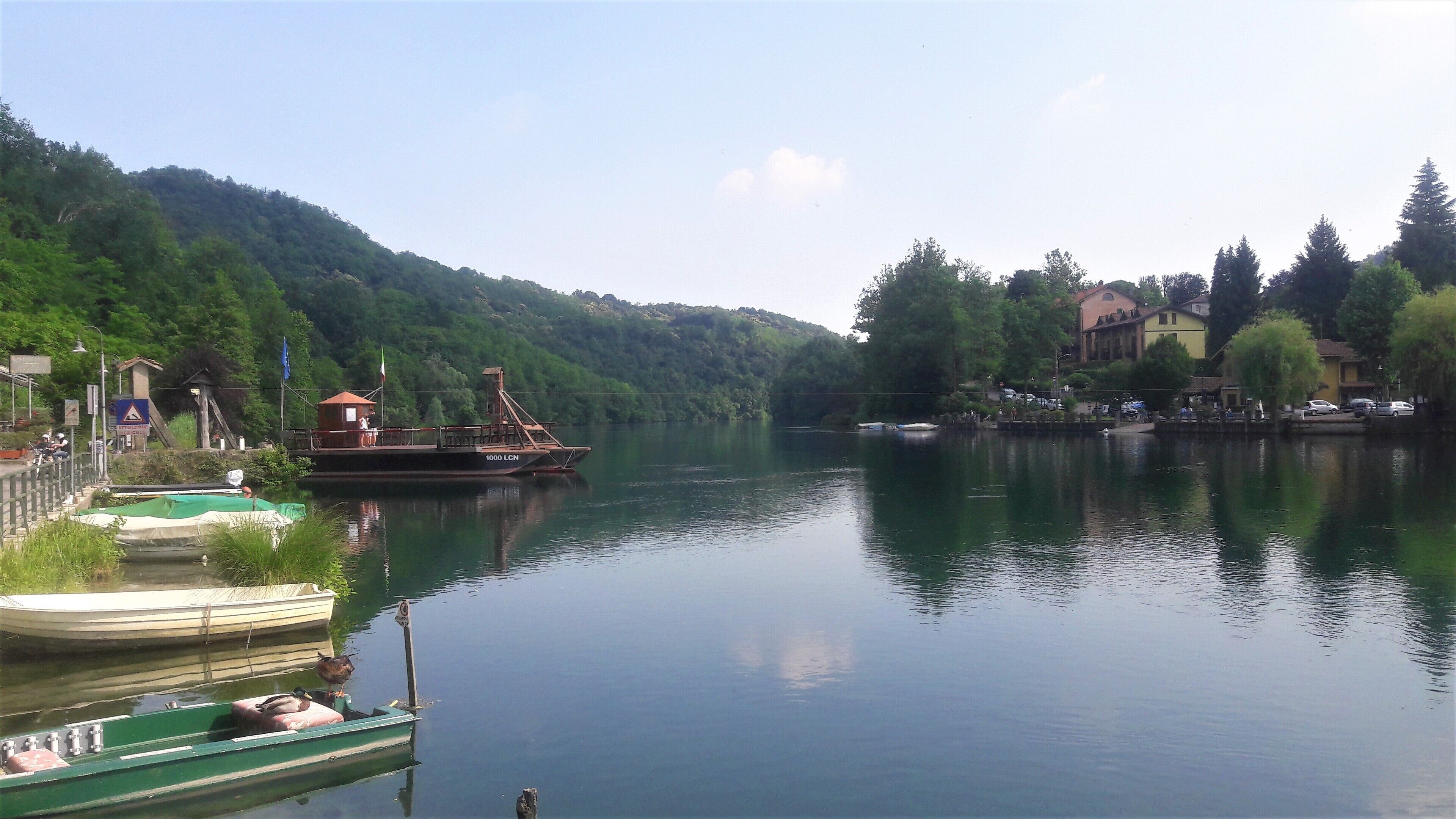 A calm river with boats docked on the left, surrounded by lush green hills and a few buildings in the background.