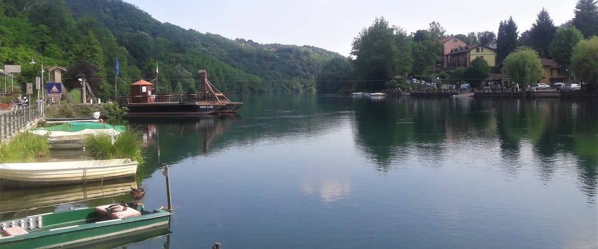 A calm river with boats docked on the left, surrounded by lush green hills and a few buildings in the background.
