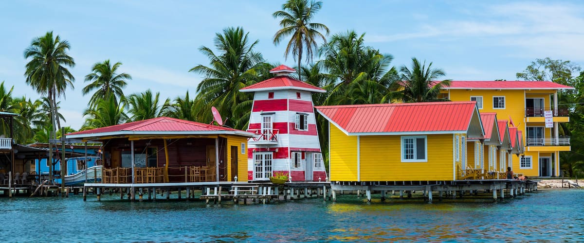 A row of colorful houses with red roofs on stilts over water, surrounded by palm trees.