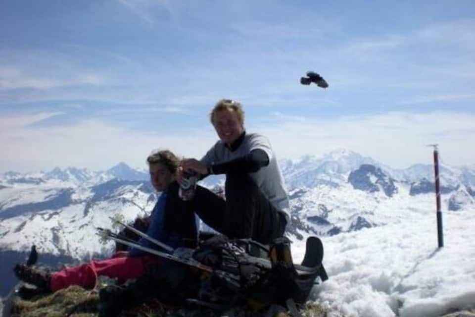 Dos personas sentadas en una montaña nevada con equipo de esquí, cielo despejado y picos nevados al fondo.