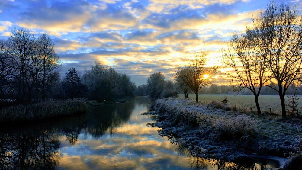 A river reflecting a sunset sky, surrounded by trees and frosty vegetation.