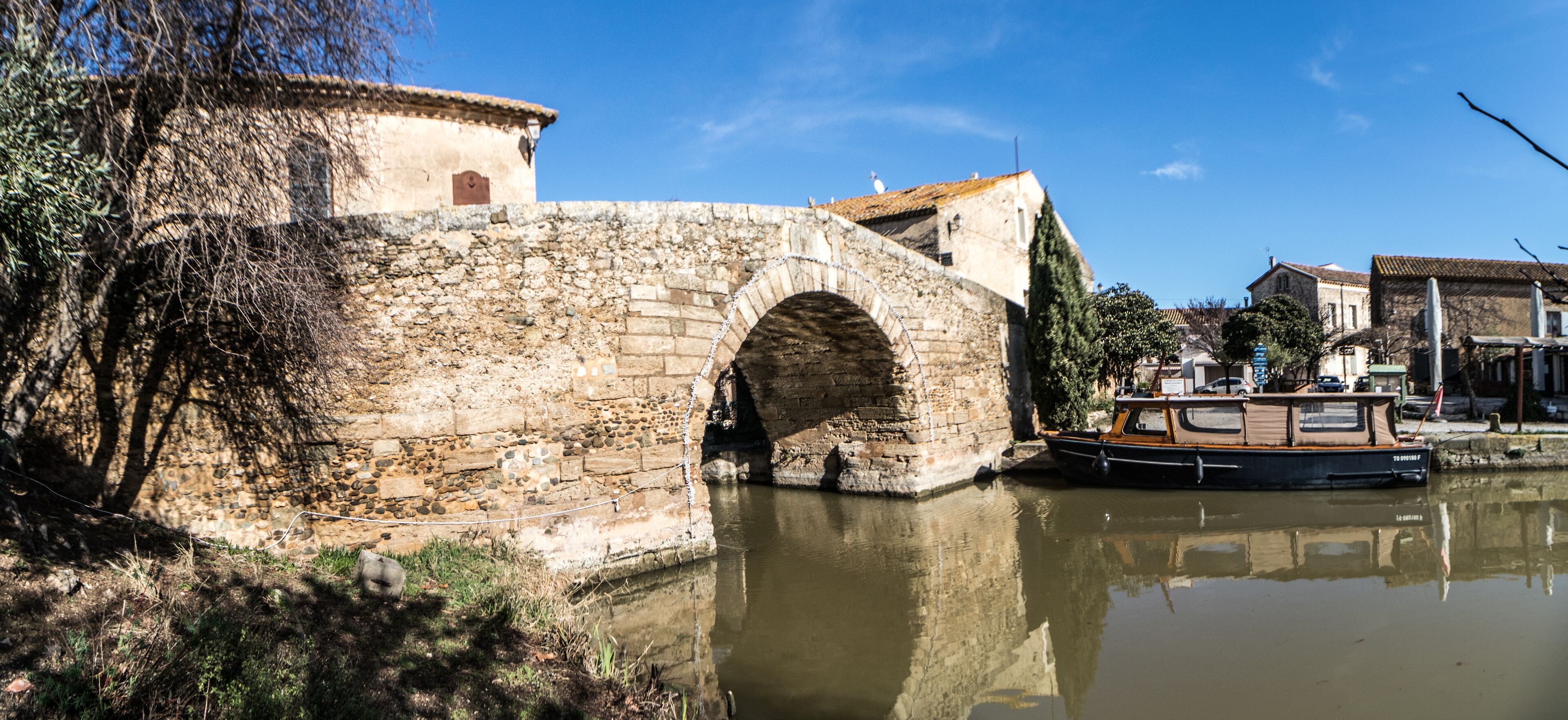 A stone bridge over a calm river, a boat docked on the right, and a building with a tiled roof in the background.