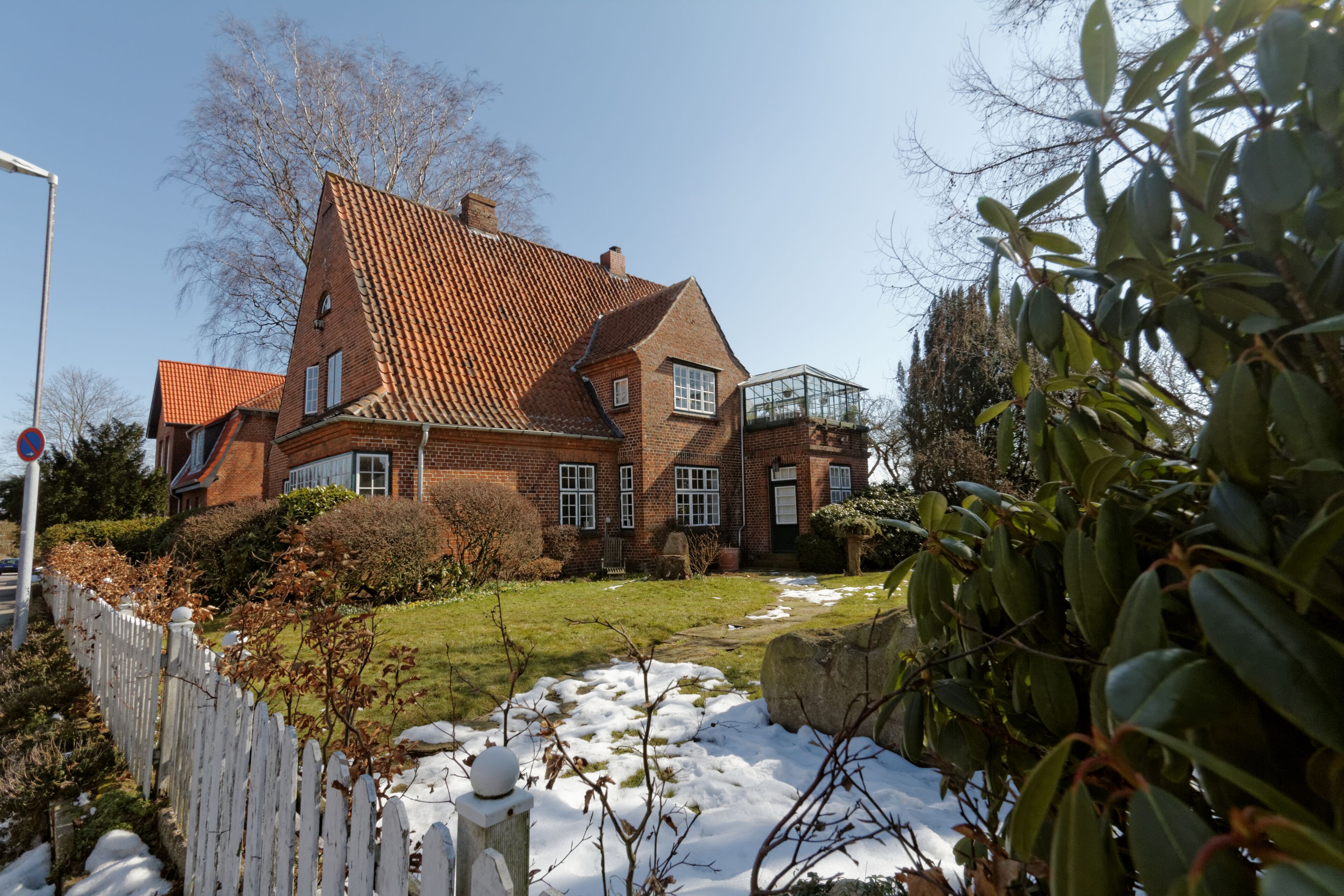 A large brick house with a sloped roof, a white picket fence, and a garden with snow patches.