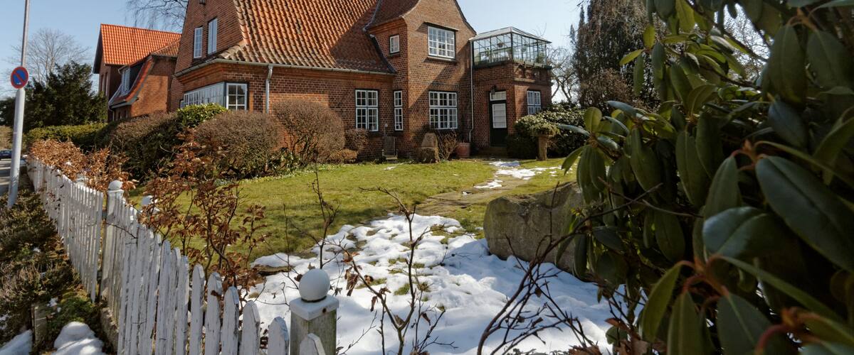 A large brick house with a sloped roof, a white picket fence, and a garden with snow patches.