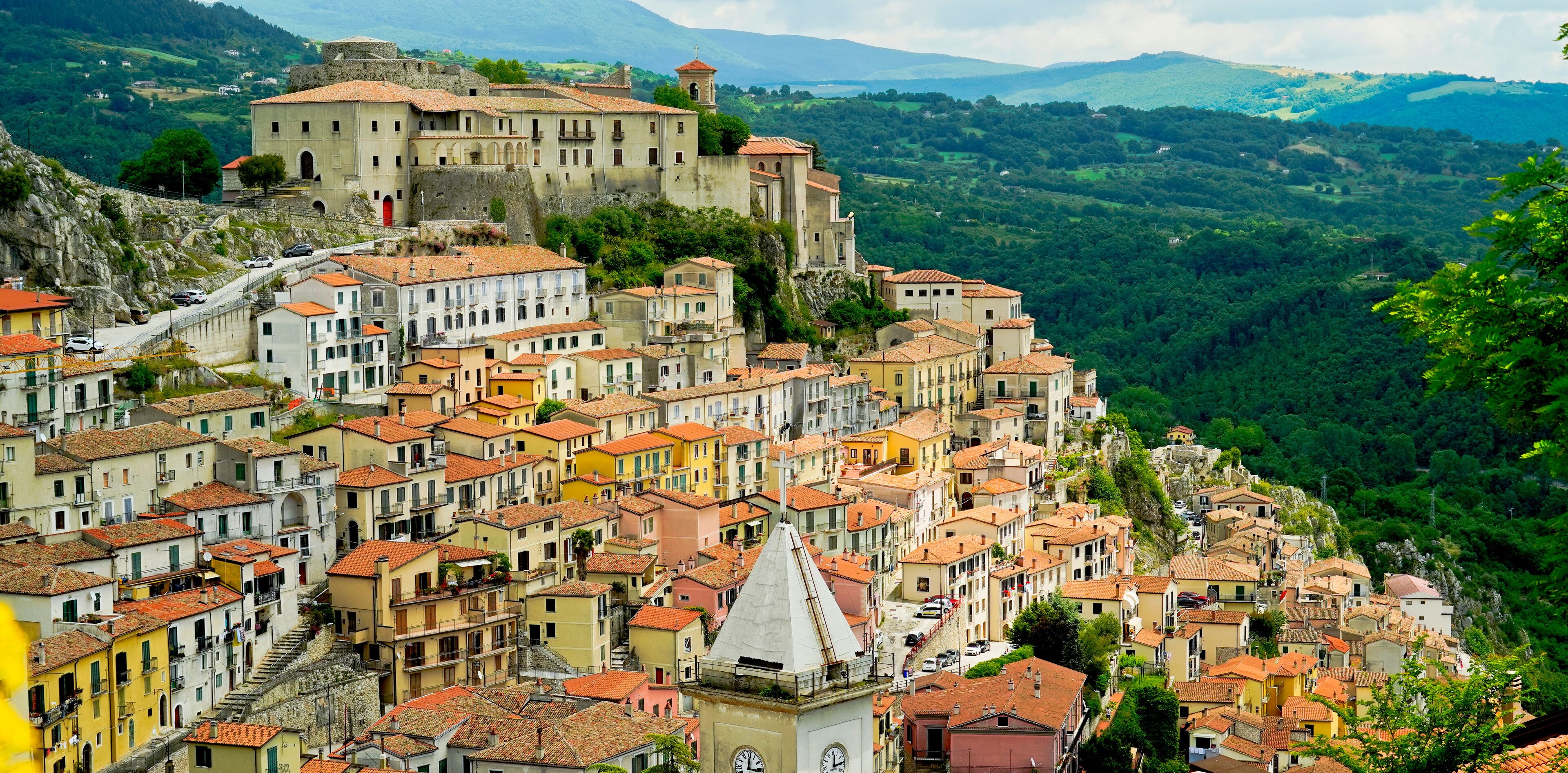 A historic town with terracotta-roofed buildings nestled in a valley, a prominent church, and a castle on a hill.
