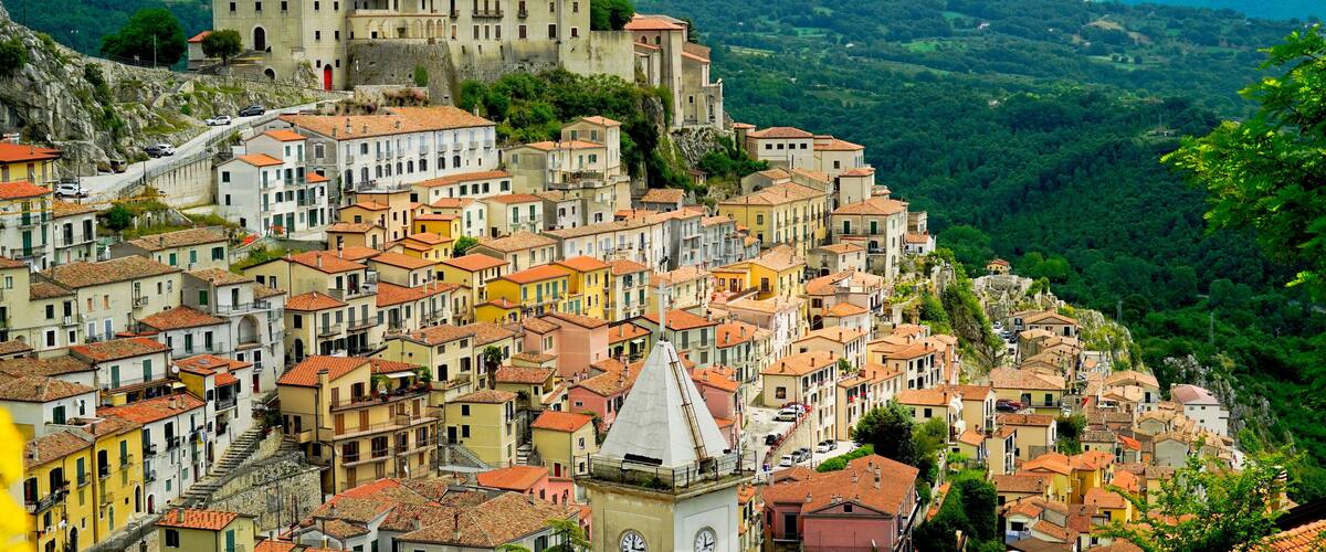 A historic town with terracotta-roofed buildings nestled in a valley, a prominent church, and a castle on a hill.