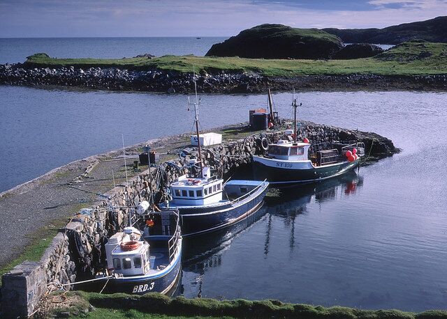 Three boats docked at a rocky pier with a calm sea and green hills in the background.
