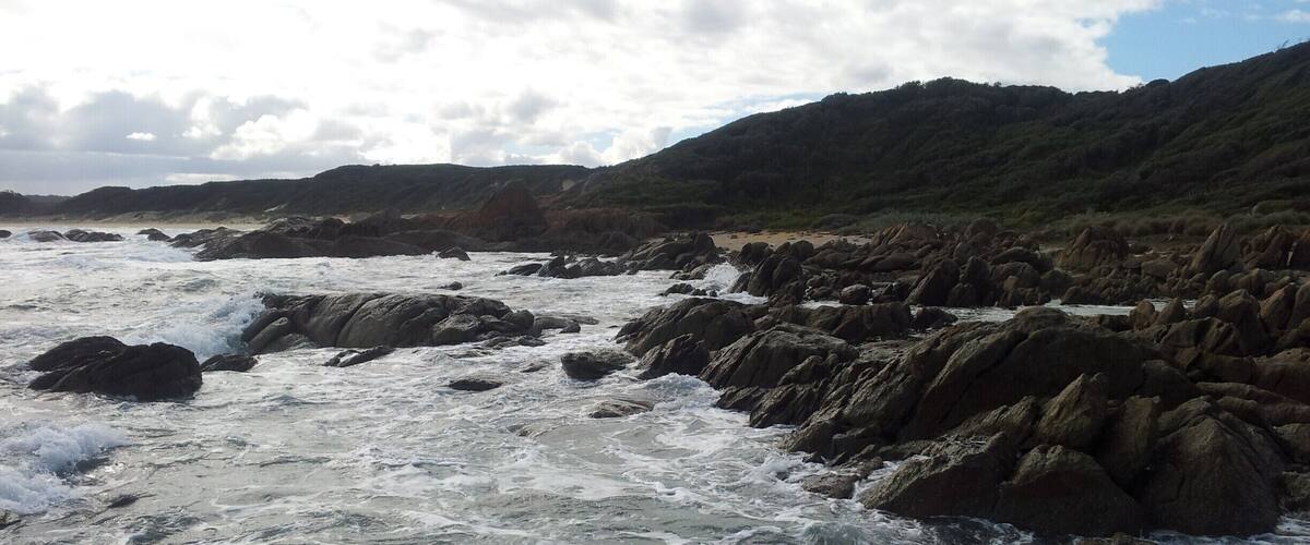 A rocky coastline with turbulent waves and a hilly landscape in the background.