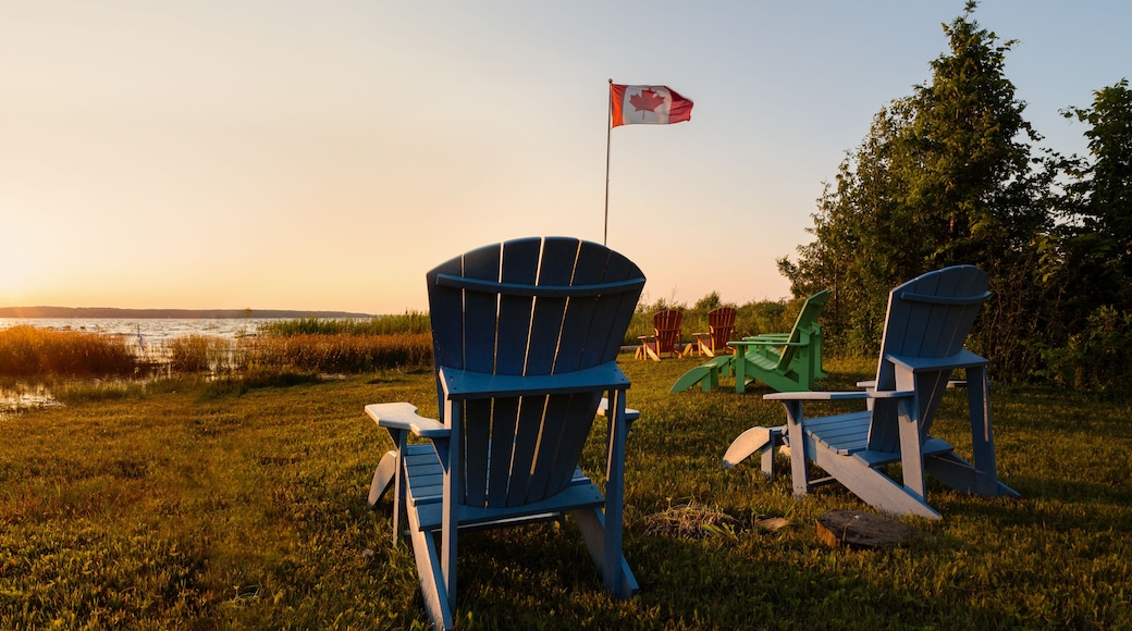 Muskoka chairs on a lawn beside a lake with a Canadian flag in the background at sunset.