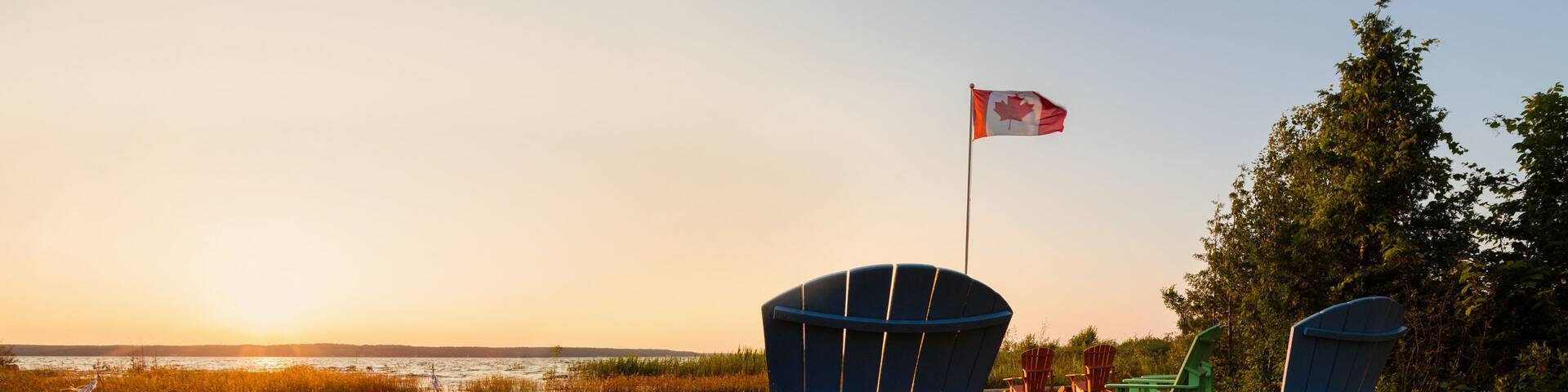 Muskoka chairs on a lawn beside a lake with a Canadian flag in the background at sunset.