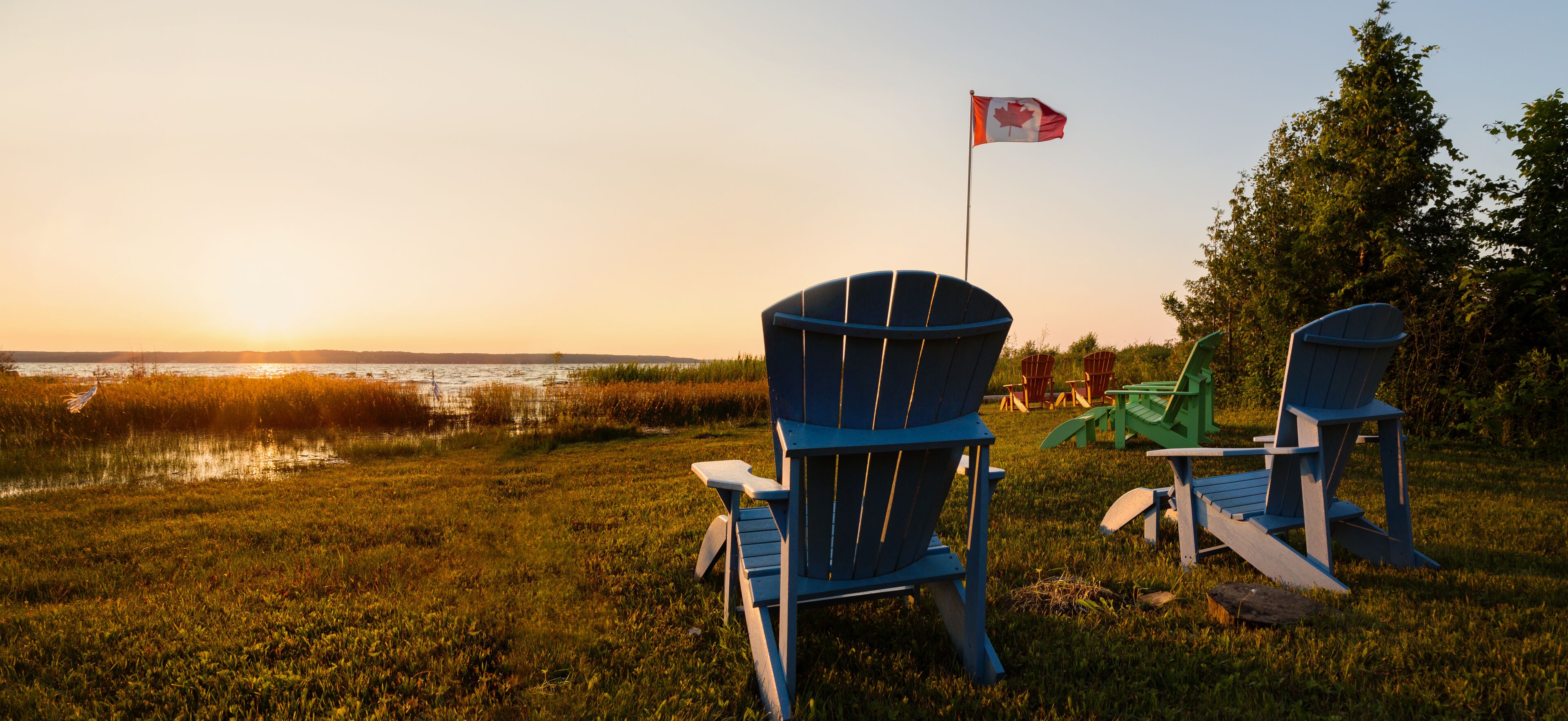 Two blue Adirondack chairs, a Canadian flag, and a sunset over a body of water.