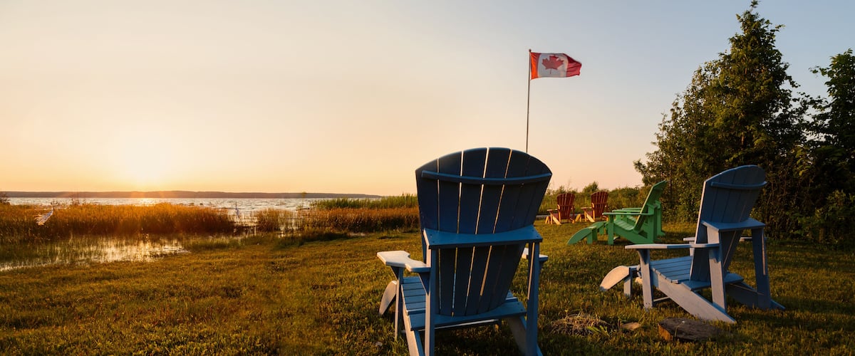 Two blue Adirondack chairs, a Canadian flag, and a sunset over a body of water.