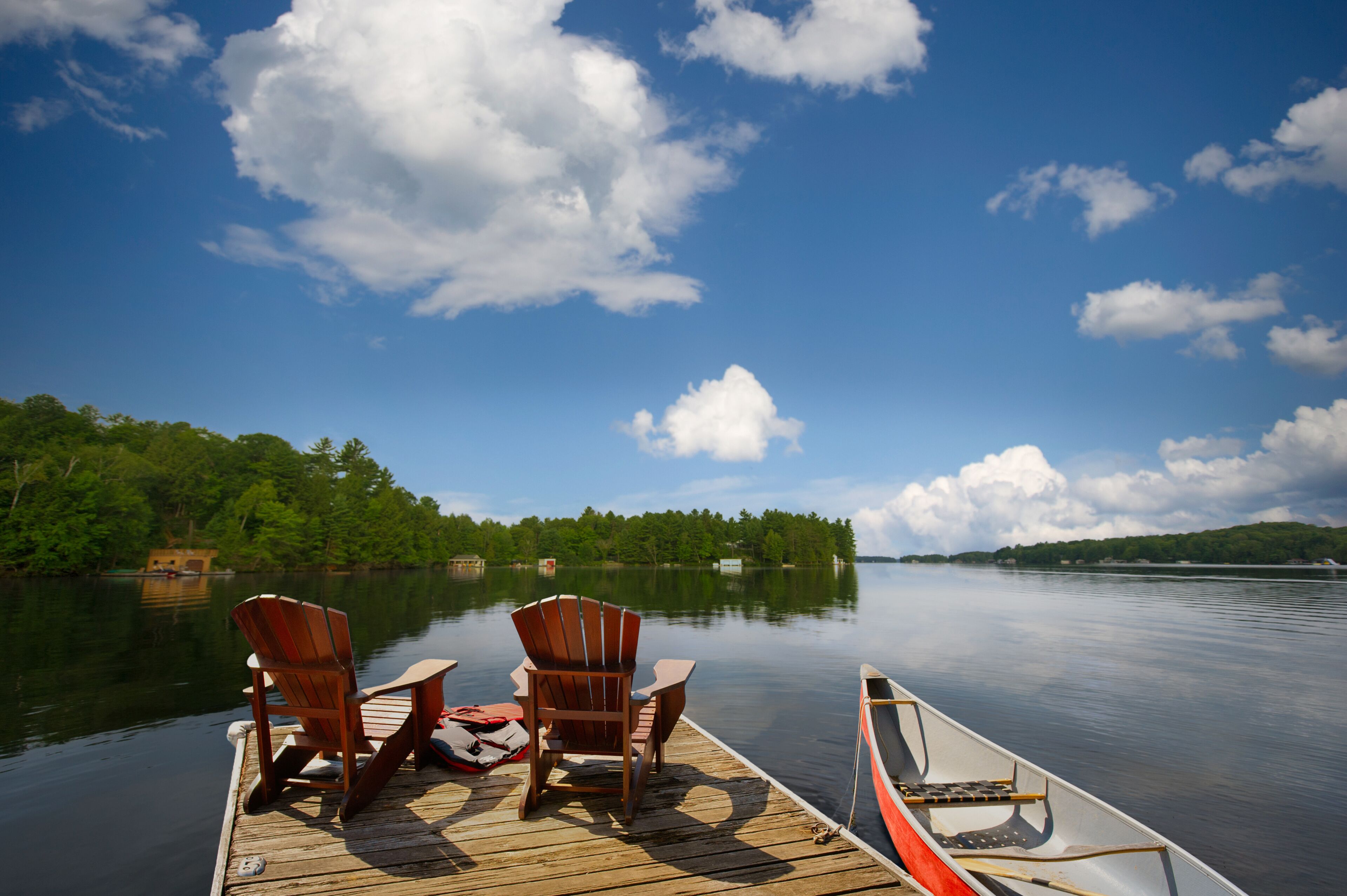 Two Adirondack chairs sit on a wooden dock overlooking a blue Muskoka lake. A canoe is tied up nearby with paddles resting inside, while fluffy white clouds drift across the summer sky.