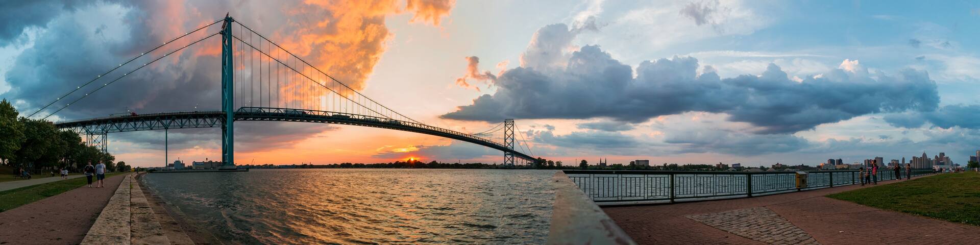 A suspension bridge over a river at sunset with a city skyline in the background.