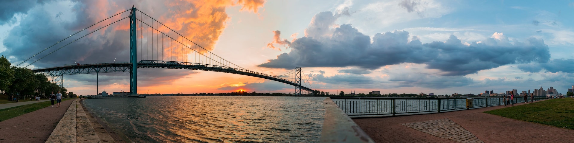 Uma ponte suspensa sobre um rio ao pôr do sol, com o horizonte da cidade ao fundo.
