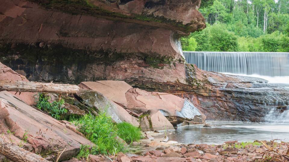 A waterfall cascading over a rocky cliff, surrounded by greenery and a calm river.