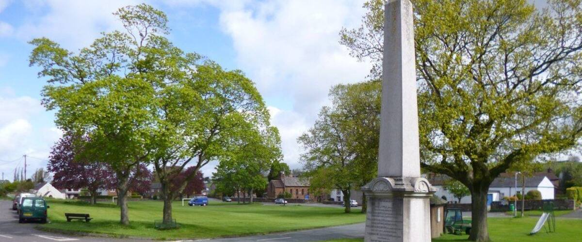 A tall monument in a grassy area with trees and a road nearby.