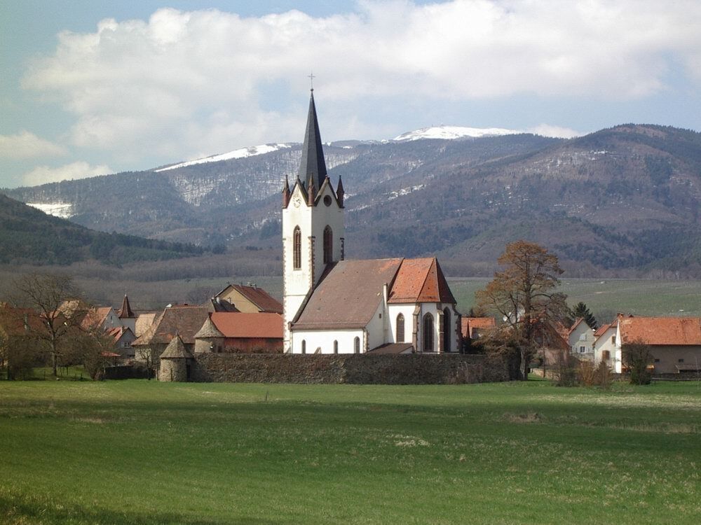 A church with a tall spire, surrounded by houses and a green field, with snow-capped mountains in the background.