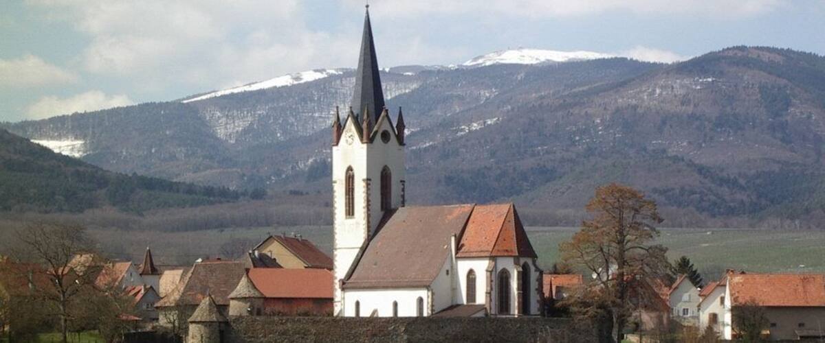 A church with a tall spire, surrounded by houses and a green field, with snow-capped mountains in the background.