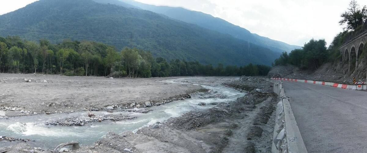 A road with a river crossing, surrounded by mountains and trees.