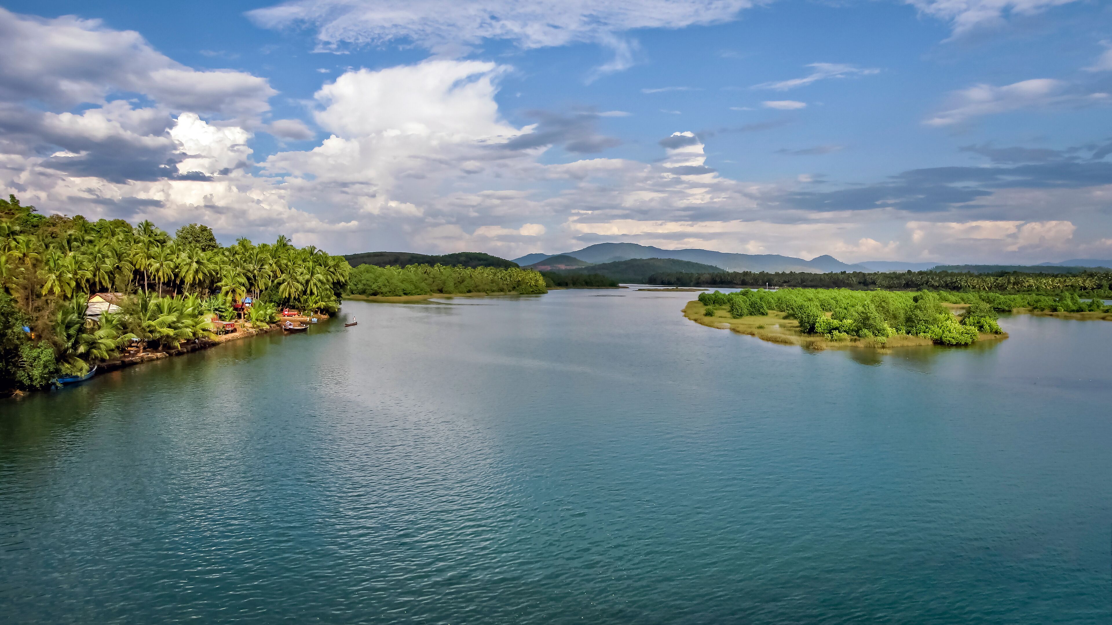 A river with dense greenery on both sides, boats, and distant mountains under a partly cloudy sky.