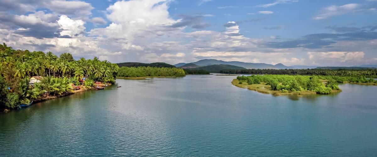 A river with dense greenery on both sides, boats, and distant mountains under a partly cloudy sky.