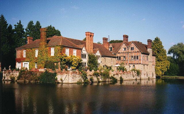 A large, historic house with multiple chimneys and ivy-covered walls, situated by a calm lake.