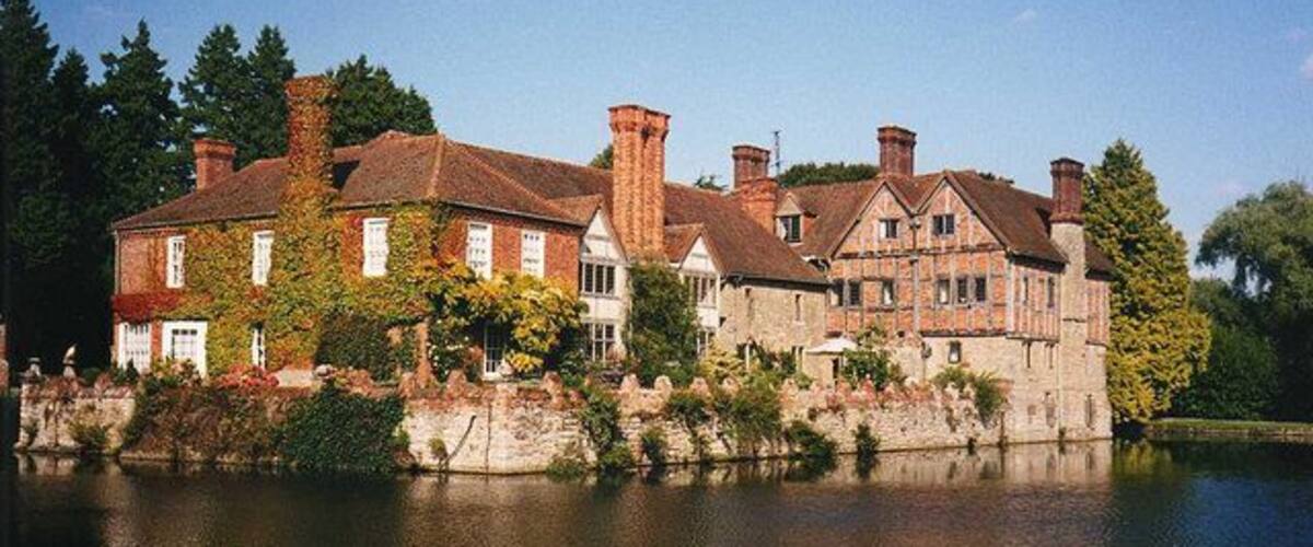 A large, historic house with multiple chimneys and ivy-covered walls, situated by a calm lake.