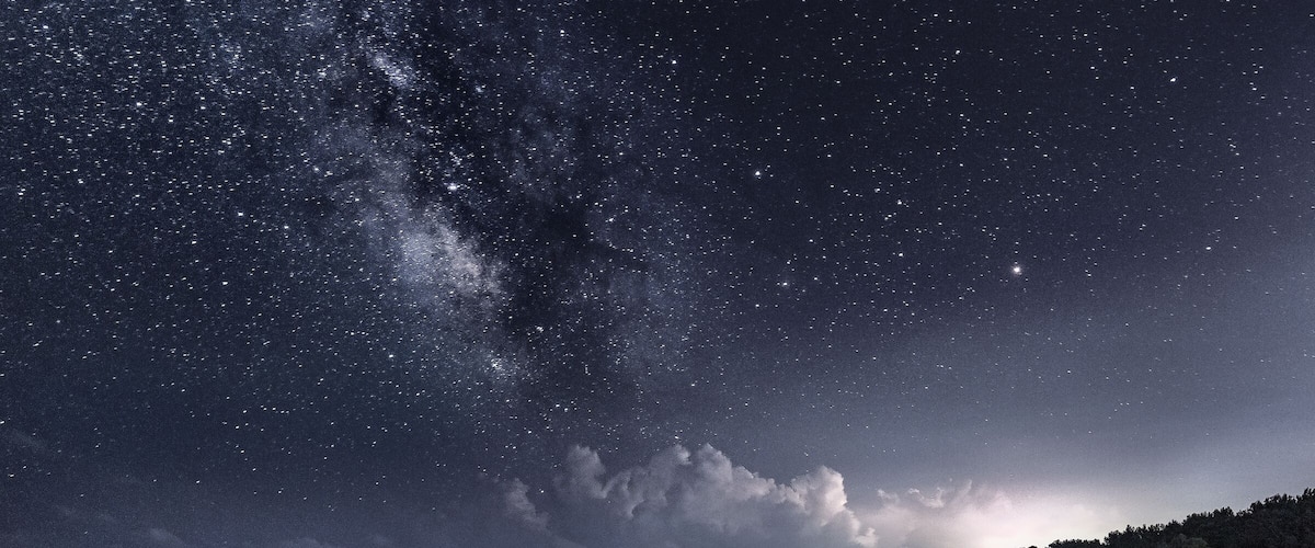 A starry night sky over a coastal landscape with a visible Milky Way and a stormy horizon.