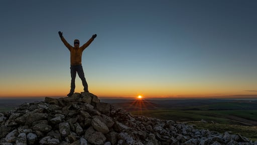 Une personne se tient debout sur un promontoire rocheux, les bras levés, et contemple le coucher de soleil sur une vallée.
