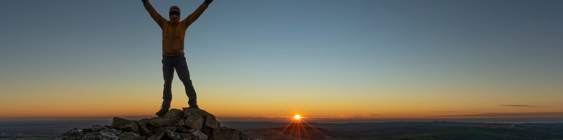 Una persona de pie sobre un saliente rocoso con los brazos levantados, contemplando una puesta de sol sobre un valle.