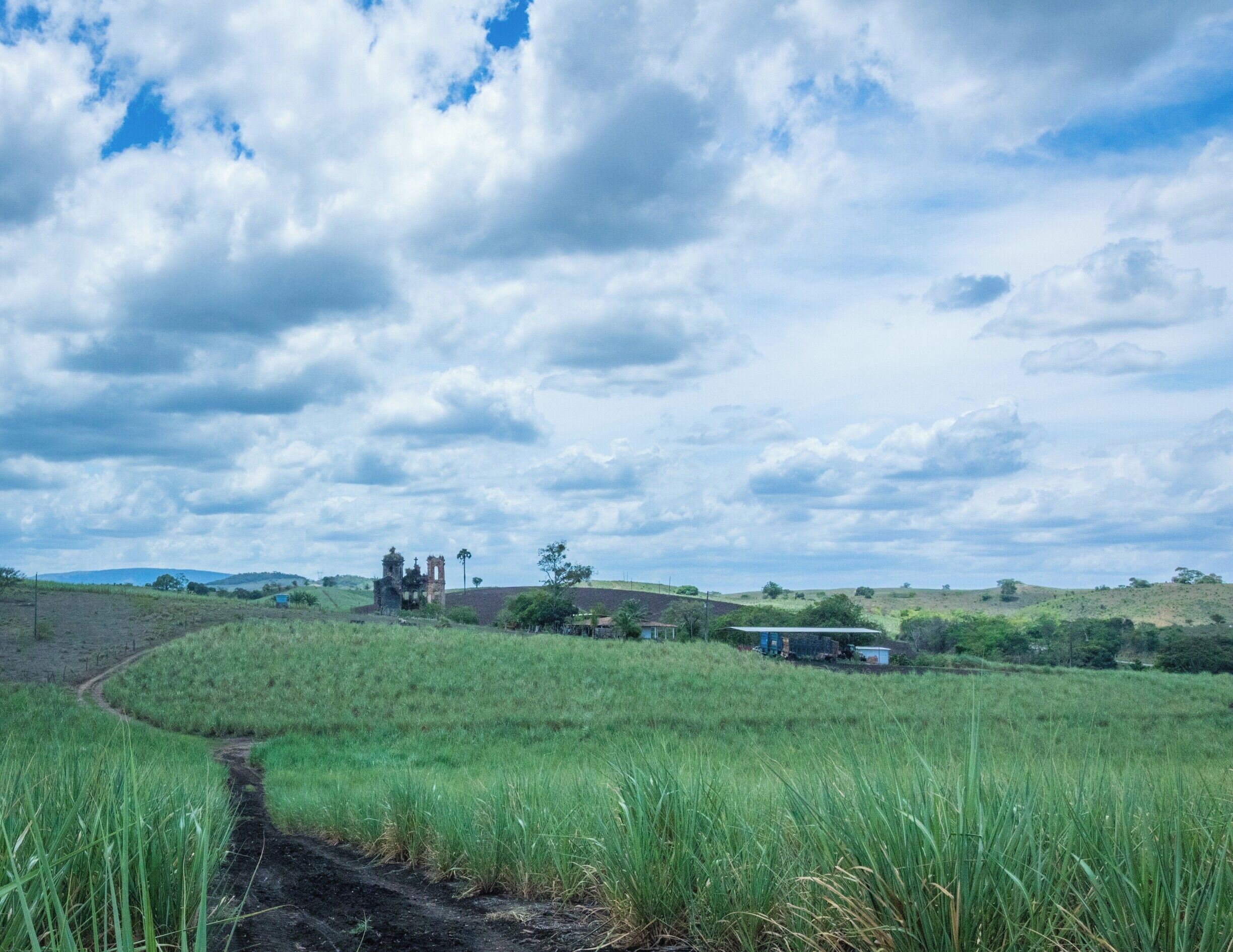 A rural landscape with a dirt path, green fields, a church, and rolling hills under a cloudy sky.