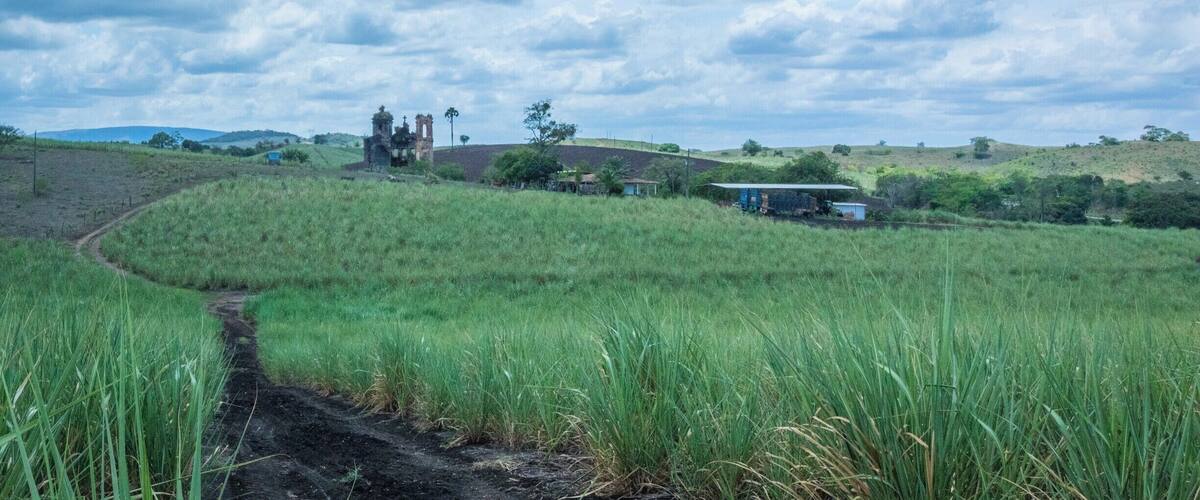 A rural landscape with a dirt path, green fields, a church, and rolling hills under a cloudy sky.