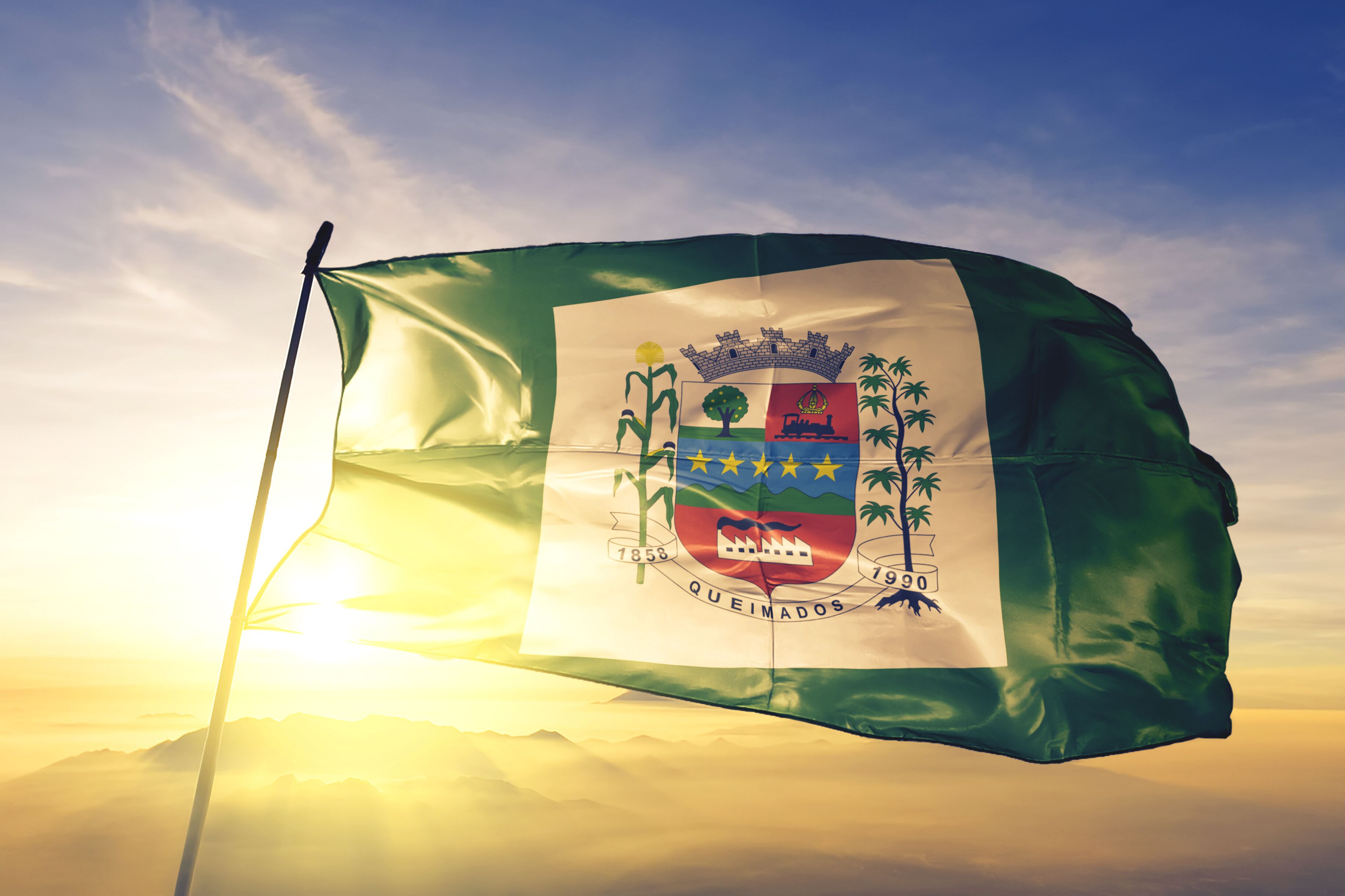 A flag with a coat of arms, the year 1853, and the word "QUEIMADOS" on a flagpole against a sunrise sky.