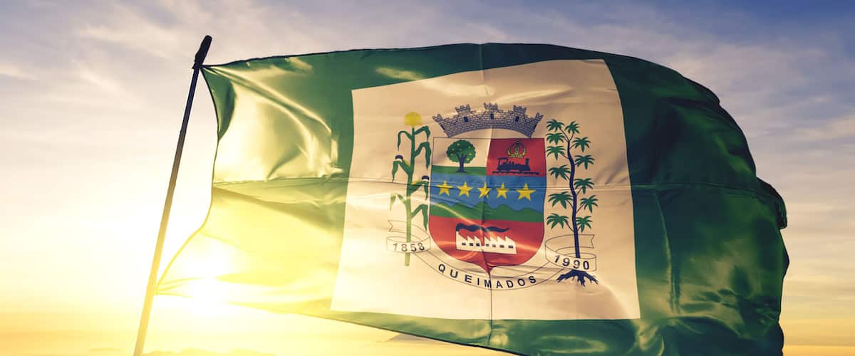 A flag with a coat of arms, the year 1853, and the word "QUEIMADOS" on a flagpole against a sunrise sky.