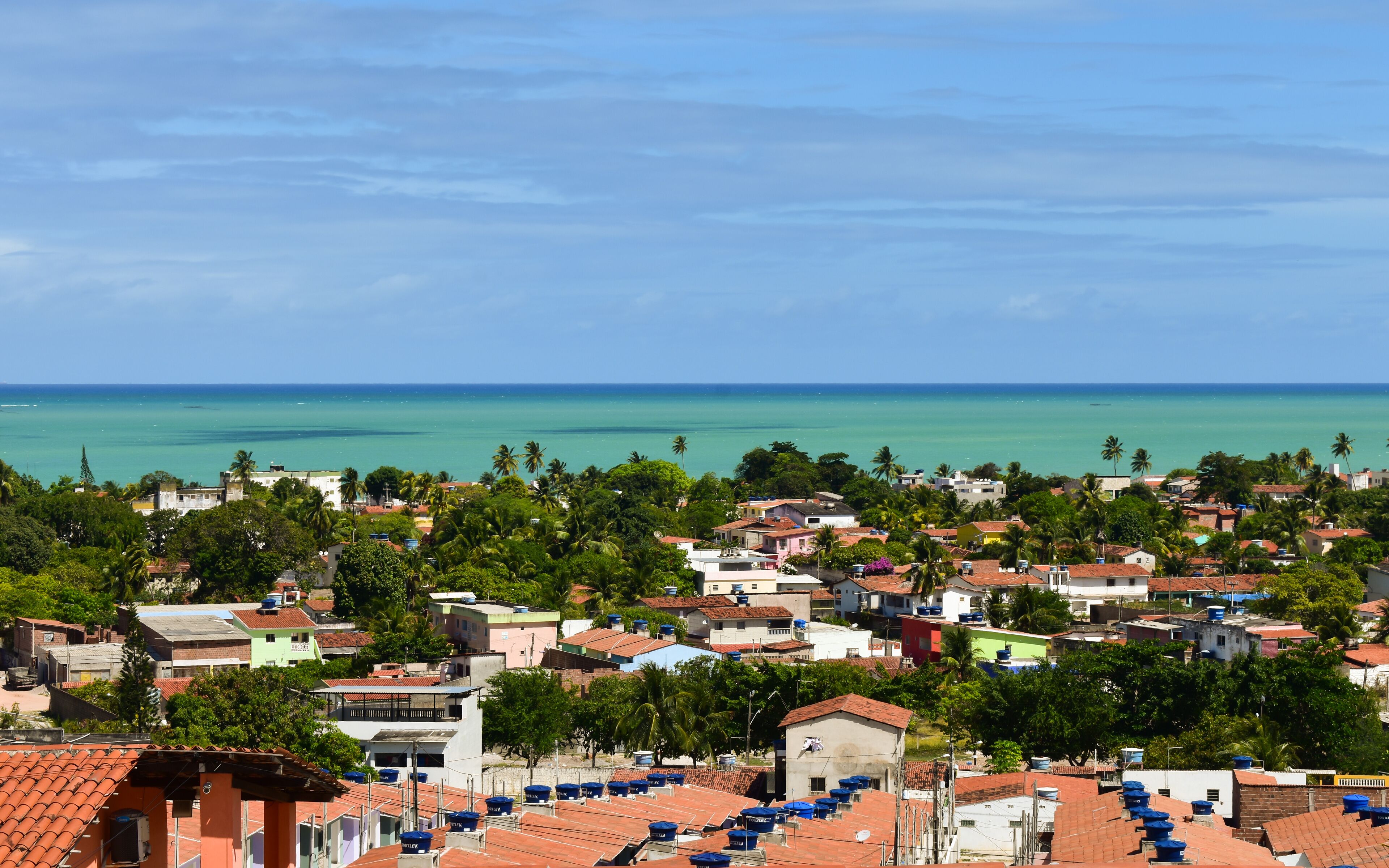 A coastal town with terracotta-roofed buildings, palm trees, and a clear view of the ocean.