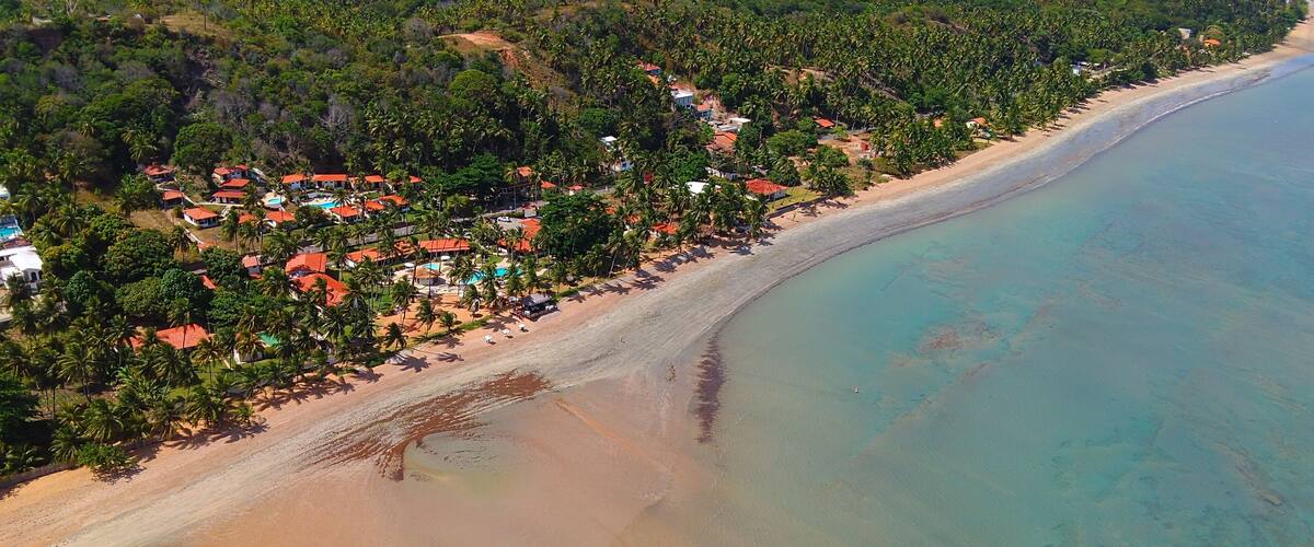 An aerial view of a beach with clear turquoise waters, a sandy shore, and a cluster of buildings with red roofs surrounded by palm trees.