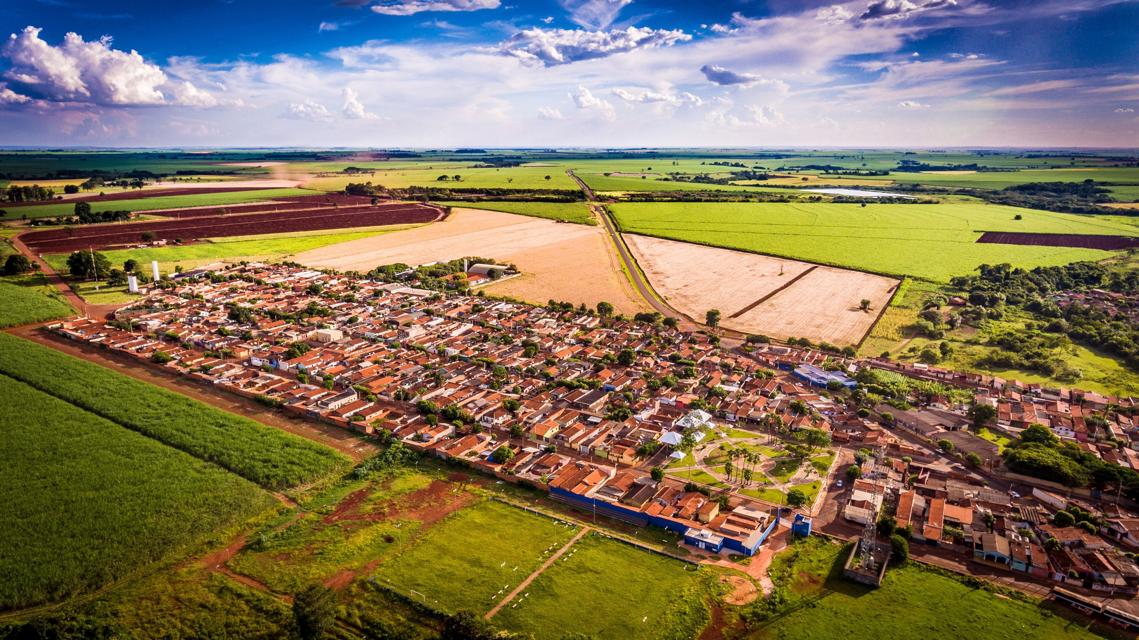 An aerial view of a town with a grid of houses, surrounded by green fields and a large open area.
